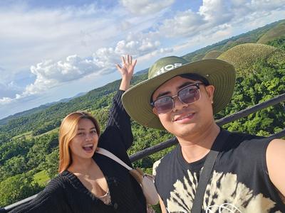 Rica Mae and Mark Lino taking a joyful selfie above green rolling hills